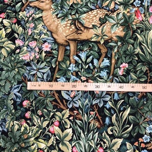 May include: A tapestry fabric featuring a dark green, brown, and blue floral pattern with two deer standing in the center. The fabric is measured with a ruler in inches.