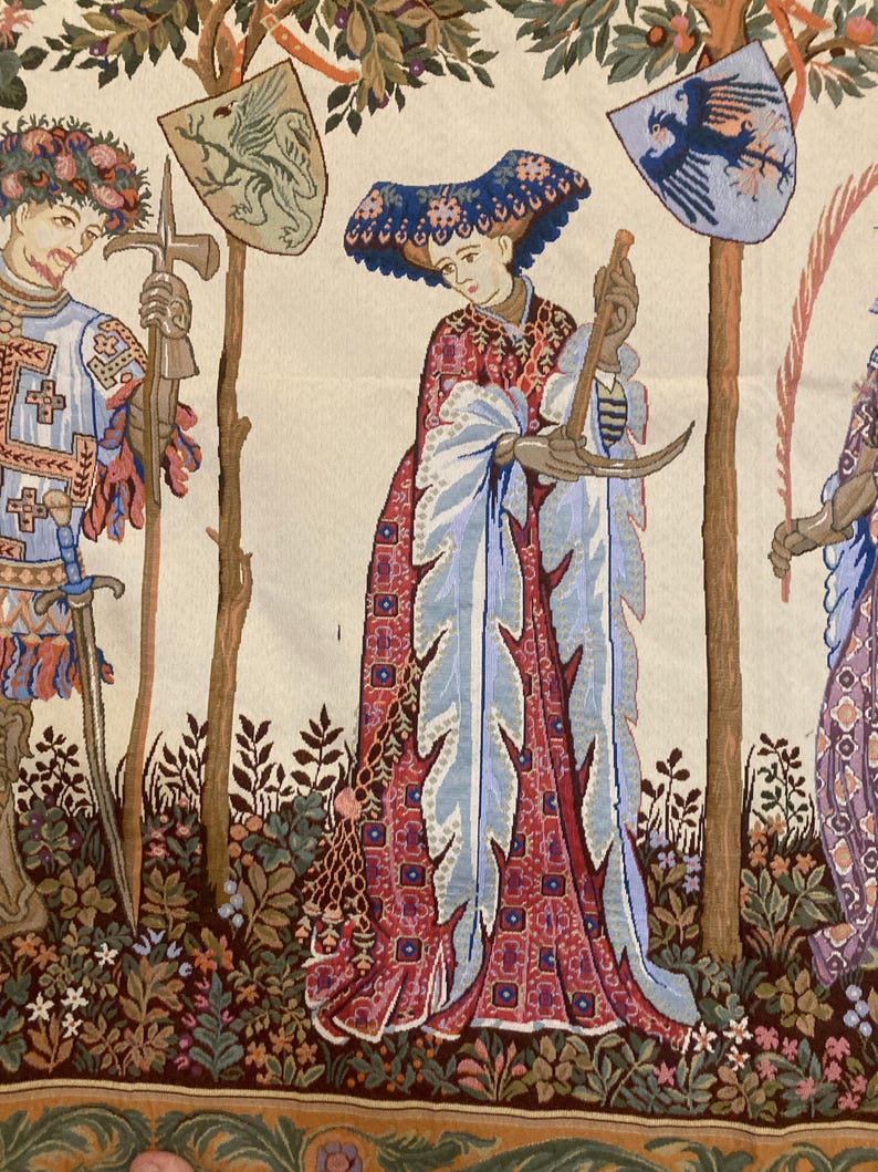 May include: A tapestry depicting a scene from medieval times. A woman in a blue and red dress holds a crescent moon in her hands. A man in armor stands to the left, holding a sword and a shield with a dragon on it. A tree with a shield featuring a bird is in the background.