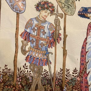 May include: A tapestry depicting a knight in armor with a sword and a shield. The knight is wearing a blue and white tunic with a red and white sash. He is standing in a forest setting with trees and flowers.