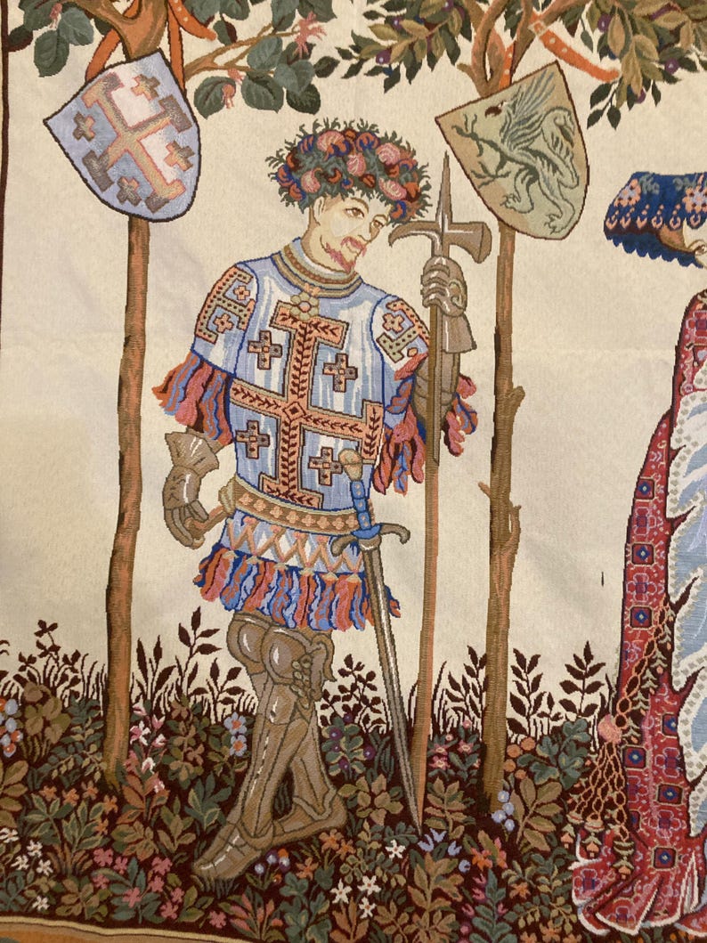 May include: A tapestry depicting a knight in armor with a sword and a shield. The knight is wearing a blue and white tunic with a red and white sash. He is standing in a forest setting with trees and flowers.