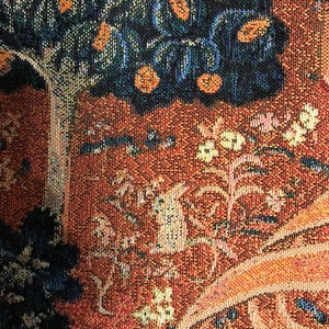 May include: A close-up of a tapestry with a brown, orange, and blue pattern. The tapestry features a tree with dark blue leaves and orange fruit, and a winding vine with white flowers. The tapestry is made of woven fabric with a textured surface.