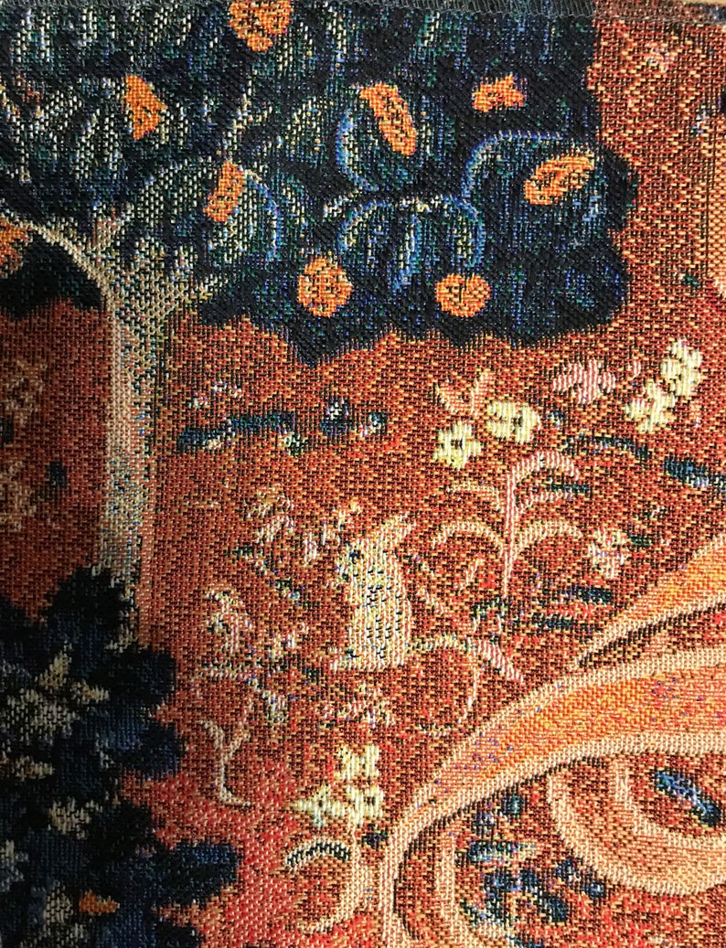 May include: A close-up of a tapestry with a brown, orange, and blue pattern. The tapestry features a tree with dark blue leaves and orange fruit, and a winding vine with white flowers. The tapestry is made of woven fabric with a textured surface.