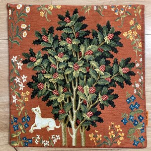 Op de afbeelding: Een kussenhoes van tapijt met een rode achtergrond met een groene boom met rode bessen en een witte hond. De boom is omringd door verschillende bloemen in tinten wit, geel, blauw en oranje.
