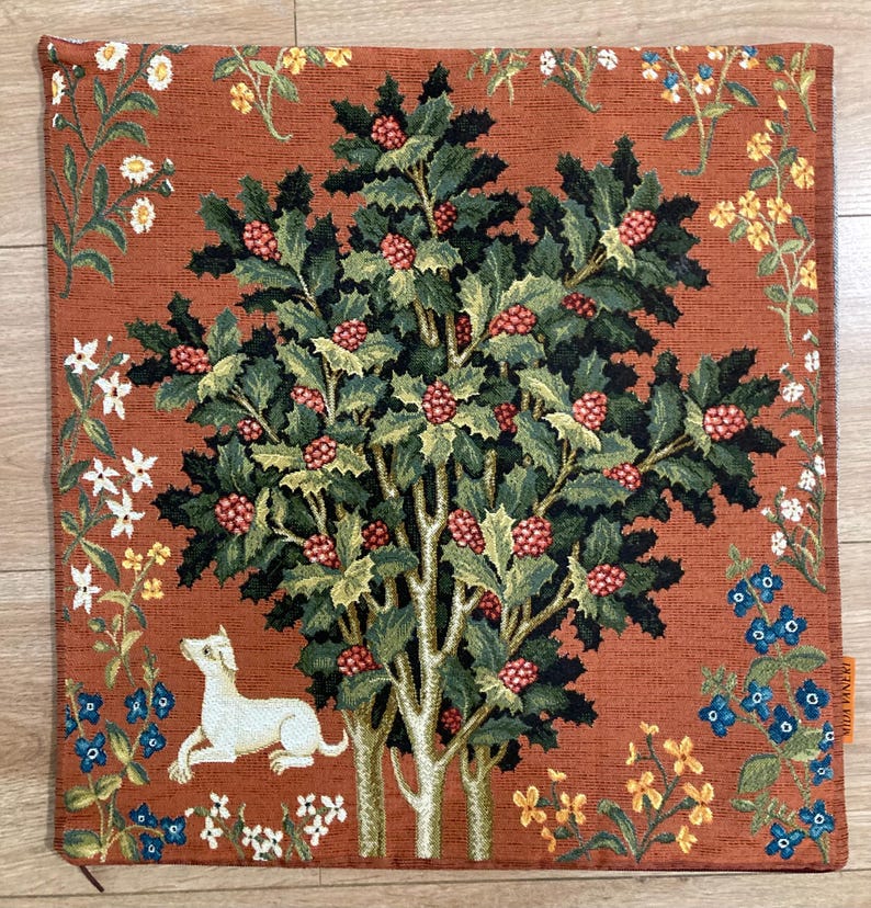 Op de afbeelding: Een kussenhoes van tapijt met een rode achtergrond met een groene boom met rode bessen en een witte hond. De boom is omringd door verschillende bloemen in tinten wit, geel, blauw en oranje.