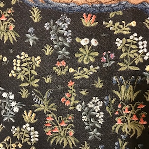 Op de afbeelding: Een tapijt met een zwarte achtergrond met een verscheidenheid aan kleurrijke bloemen en planten. Het tapijt heeft een blauwe rand en een bruin en blauw gedeelte bovenaan.