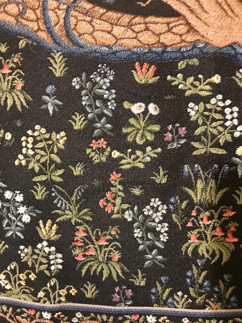 Op de afbeelding: Een tapijt met een zwarte achtergrond met een verscheidenheid aan kleurrijke bloemen en planten. Het tapijt heeft een blauwe rand en een bruin en blauw gedeelte bovenaan.