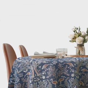 May include: A round, navy blue tablecloth with a detailed botanical and bird pattern. The design features trees, birds, and floral elements in shades of beige, green, and brown. The tablecloth is set on a table with two brown chairs.