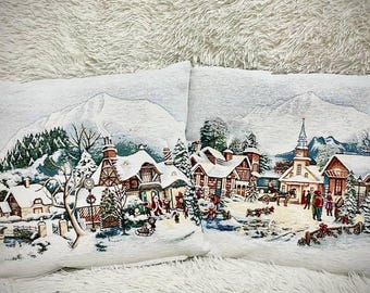 以下が含まれることがあります： 冬の村のシーンが描かれた装飾的なクッション2つ。クッションには、雪景色に家、木々、教会が描かれています。このシーンは、伝統的なクリスマスの村を彷彿とさせます。
