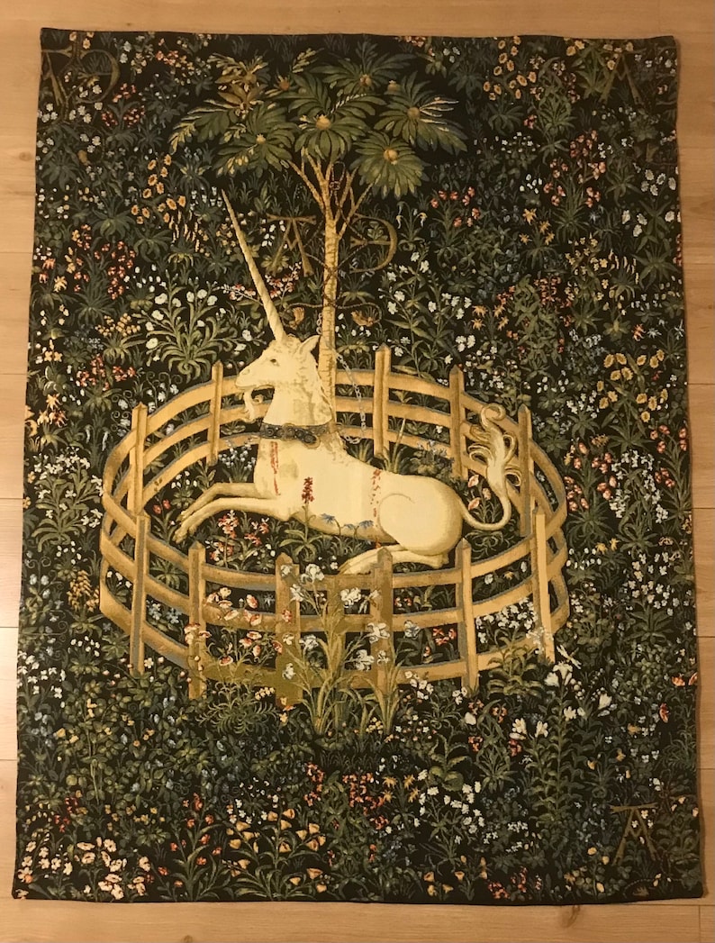 Puede incluir: Un tapiz que representa un unicornio blanco acostado en una valla circular de madera, rodeado de un exuberante bosque verde con flores de colores. El unicornio tiene un collar de oro y un cuerno.