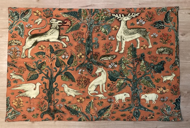 May include: A tapestry featuring a medieval-style design with a lion, deer, unicorn, and other animals on a red-orange background. The tapestry is woven with intricate details and features a variety of green and brown foliage.