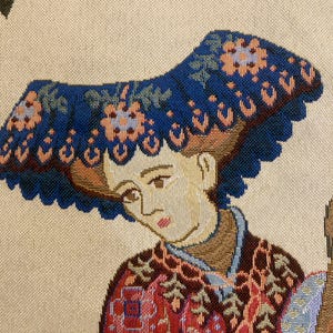 May include: A tapestry featuring a woman wearing a blue hat with floral embroidery. She is wearing a red and gold patterned dress. The tapestry has a cream background.
