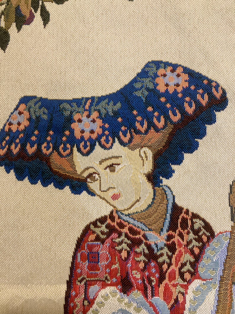 May include: A tapestry featuring a woman wearing a blue hat with floral embroidery. She is wearing a red and gold patterned dress. The tapestry has a cream background.