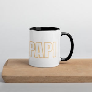 Może przedstawiać: Biały kubek ceramiczny z czarnym rantem i uchwytem. Kubek ma napis "PAPI" nadrukowany na przodzie pomarańczowym kolorem.