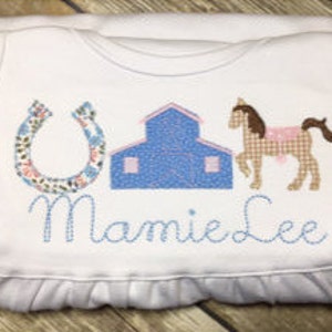 May include: White baby onesie with a blue barn, brown horse with a pink saddle, and a horseshoe with floral accents. The onesie is personalized with the name "Mamie Lee".