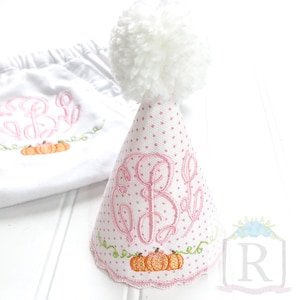 以下が含まれることがあります： 白とピンクのドット柄のパーティーハットで、トップに白いポンポンが付いています。ハットにはピンクの刺繍のモノグラムと、オレンジ色のカボチャと緑色のツルがデザインされています。