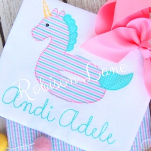 May include: White t-shirt with a pink and blue striped unicorn float design. The shirt is personalized with the text "Robinson Lane Andi Adele".