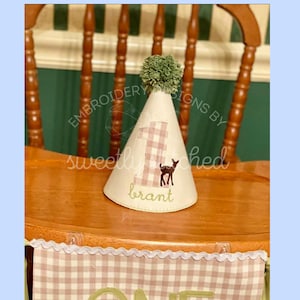 May include: A pink and white gingham fabric party hat with a green pom-pom on top. The hat has a number one and a deer embroidered on it. The hat says "brant" on the bottom.