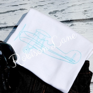 May include: White t-shirt with a blue outline of a vintage airplane. The text "Robinson Lane" is embroidered on the shirt.