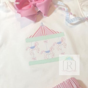 May include: A white embroidered shirt with a pink and green carousel design. The carousel has three horses and a pink and white striped roof.