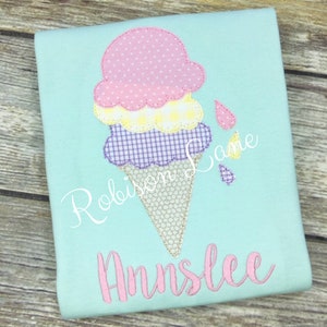 May include: A light blue t-shirt with a pink, yellow, and purple ice cream cone applique. The ice cream cone has a brown waffle cone and three scoops of ice cream. The ice cream is melting and dripping down the side of the cone. The shirt also has the name "Annslee" embroidered in pink on the front.