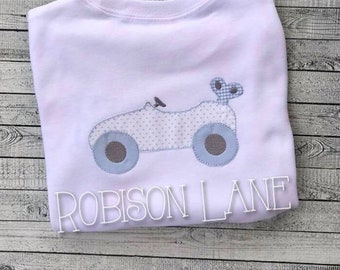 Puede incluir: Una camiseta blanca de manga larga con un aplique de tractor azul claro y gris. El tractor tiene detalles de lunares y ruedas azules. El texto "ROBISON LANE" está impreso en la parte inferior de la camiseta.