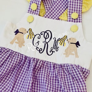 May include: A white and purple gingham baby overall with yellow buttons and a yellow bow. The overall has an embroidered monogram "R" and two embroidered yellow dogs with yellow bows.