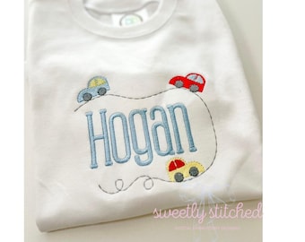 Puede incluir: Camiseta blanca con un diseño bordado. El nombre "Hogan" en azul claro está rodeado por una línea punteada y tres coches de juguete de colores: azul, rojo y amarillo.