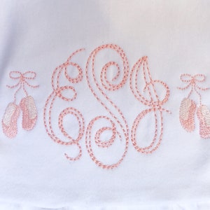 May include: White fabric with pink embroidered ballerina shoes and a monogrammed letter "S" in a cursive script.