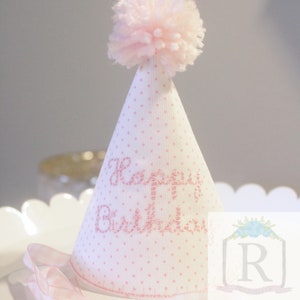 May include: A white party hat with pink polka dots and a pink pom-pom on top. The hat says "Happy Birthday" in pink glitter.