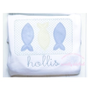 Puede incluir: Un pelele blanco con un diseño bordado de tres peces azules y amarillos, con el nombre "hollis" cosido debajo. El diseño está rodeado por un borde de puntos.