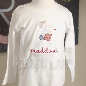 May include: White long-sleeve shirt with an embroidered image of a boy playing a drum. The shirt has the name "maddox" and the text "ROBISSON LANE" embroidered on it.