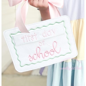 May include: A white felt sign with a pink ribbon handle. The sign says "first day of school" in pink embroidery.