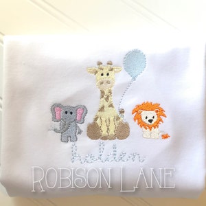 May include: White embroidered baby onesie with a giraffe, elephant, and lion. The onesie has the name "Holden Robison Lane" embroidered on it.