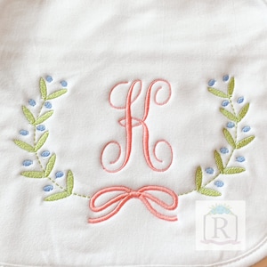 May include: A white embroidered bib with a pink letter "K" surrounded by a green wreath with blue berries and a pink bow.