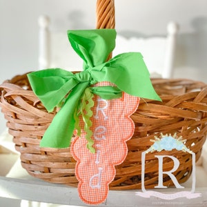 May include: A woven wicker basket with a green fabric bow and a carrot-shaped tag. The tag is orange gingham with the name "Reid" embroidered in white. The basket has a woven handle.