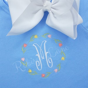 May include: Light blue shirt with a white bow and embroidered monogram in a floral wreath. The monogram is "M" with the name "Robinson" written below it.