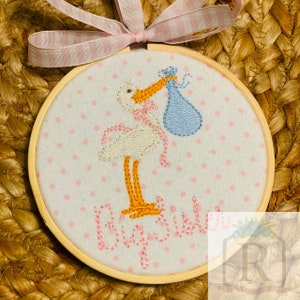 May include: A white stork with orange legs and a pink ribbon around its neck is carrying a blue baby bundle. The stork is embroidered on a white fabric with pink polka dots. The words "Big Sis" are embroidered in pink below the stork. The fabric is mounted in a wooden hoop.