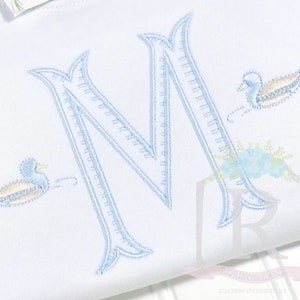 May include: A white embroidered baby onesie with a blue letter M and two blue ducks. The ducks are embroidered with yellow accents.