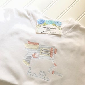 May include: White t-shirt with embroidered design of a toy gun and the name "Hollie".