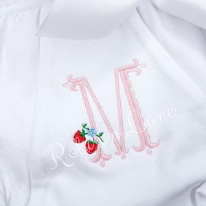 Pode incluir: Um macacão branco bordado com uma letra M rosa e dois morangos vermelhos com folhas verdes e uma flor azul.