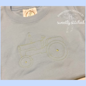 May include: A light blue t-shirt with a gold embroidered tractor design. The design includes the text "sweetly stitched" in a gold font.