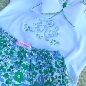 May include: A white baby onesie with a green bow and embroidered monogram "KR". The onesie is paired with a green and blue floral print bloomer with the text "ROBISSON LANE" on the bottom.