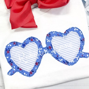 May include: A white shirt with a blue and red heart-shaped sunglasses applique. The sunglasses have a white and blue striped fabric inside the heart shape. The text "Rebel" is embroidered on the left heart and "Love" is embroidered on the right heart.