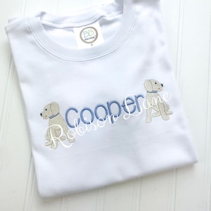 Può includere: Maglietta bianca con il testo ricamato in blu "Cooper Robinson Lane" e due cani labrador gialli ricamati.