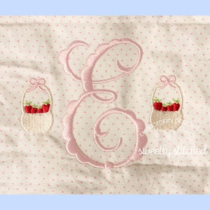Könnte beinhalten: Rosa bestickter Buchstabe E mit zwei kleinen weißen Körben gefüllt mit roten Erdbeeren. Der Text "Embroidery Designs by Sweetly Stitched" ist unter den Körben sichtbar.