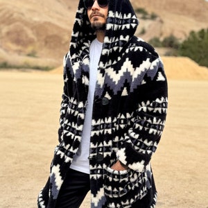 以下が含まれることがあります： 黒、白、グレーの幾何学模様のフード付きロングコート。 コートには大きなポケットがあり、ボタンで留められています。 黒いズボンとサングラスを着用しています。 背景は砂漠の風景です。