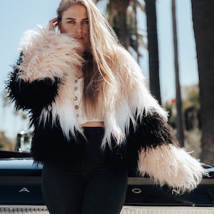 Può includere: Una donna indossa una giacca di pelliccia sintetica nera, bianca e rosa e si trova davanti a un'auto nera. L'auto ha un paraurti cromato e una finitura cromata attorno ai fanali posteriori. I fanali posteriori sono rossi e l'auto ha la scritta "Sprint" sul retro.