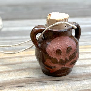 May include: A small brown ceramic bottle with a cork stopper and a painted skull and crossbones design. The bottle is hanging from a twine cord.