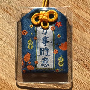 May include: A blue and white Japanese charm with a yellow string. The charm has a white panel with the text "力 事 胜 意" in black. The charm is surrounded by a blue fabric with orange and yellow flowers and other designs.