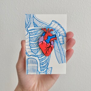 May include: A colorful anatomical heart pin, featuring red, blue, pink, and purple hues, is displayed on a white card with blue skeletal illustrations. The pin is a detailed representation of a human heart.
